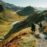 Deux randonneurs avancent sur un sentier de montagne, profitant des paysages spectaculaires. Les couleurs d'automne ajoutent une touche vibrante à la scène, tandis que les fiz s'élèvent au loin.