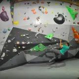 Un homme en t-shirt vert fait de l'escalade pertuis sur un mur coloré, un autre homme le regarde.