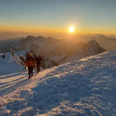 Deux alpinistes progressent sur un sentier enneigé au lever du soleil, dominant un paysage de montagnes spectaculaire. Le sommet montagneux est baigné d'une lumière dorée.