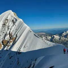 Deux alpinistes escaladent la crête enneigée de l'Aiguille Verte, sous un ciel bleu intense. Le soleil projette de longues ombres sur la montagne.