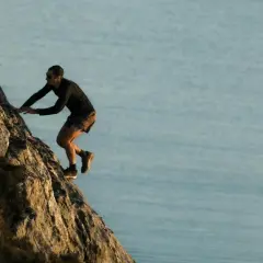 Kilian Jornet escalade une paroi rocheuse abrupte, dominant un plan d'eau calme, rappelant l'esprit d'aventure dans les Pyrénées.