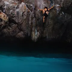 Une femme pratique l'escalade outdoor au-dessus d'une eau turquoise. Elle s'agrippe à la paroi rocheuse, concentrée sur sa prochaine prise.
