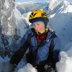 Une alpiniste souriante, Nives Meroi, en plein effort sur une crête enneigée, casque jaune, montagnes majestueuses en arrière-plan.