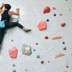 Grimpeuse en action, un pied dans la main, sur un mur d'escalade coloré. Elle progresse avec agilité.