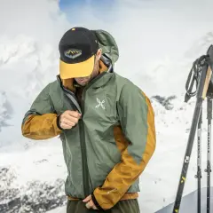 Un skieur ajuste sa veste technique, peut-être pour comparer le confort entre Primaloft vs Gore-Tex, avec des montagnes enneigées en arrière-plan.