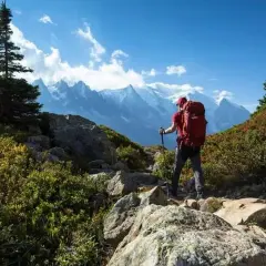 Un randonneur solo, sac à dos rouge, bâtons de marche, admire les montagnes majestueuses sous un ciel bleu éclatant.