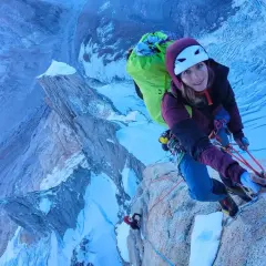 Lise Billon, casque blanc, grimpeuse souriante, est en pleine ascension sur une paroi rocheuse glacée, avec un sac à dos vert vif.
