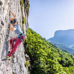 Une grimpeuse évolue sur une paroi rocheuse, profitant d'une journée ensoleillée dans ce parc d'escalade naturel. Le paysage montagneux verdoyant offre un panorama spectaculaire.