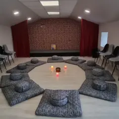 Salle de méditation avec coussins disposés en cercle et bougies allumées. Des chaises attendent autour, créant une atmosphère paisible, comme un refuge au petit goulet.