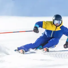 Un skieur en combinaison jaune et bleue vire sur la neige, ses skis paraboliques taillant la piste.