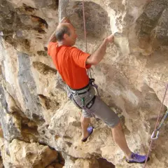 Un homme en t-shirt orange et short gris fait de l'escalade sur une paroi rocheuse. Il est assuré par une corde et semble atteindre un point élevé, un vrai **grimp azur**.