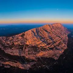 Le soleil couchant illumine la montagne Sainte-Victoire, offrant un spectacle grandiose pour les amateurs d'escalade. La lune veille sur ce paysage rocheux.
