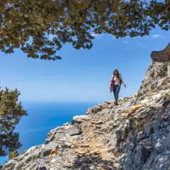 Une randonneuse explore un sentier rocailleux sur une montagne de Crète, avec la mer bleue en arrière-plan.