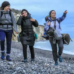 Trois randonneuses marchent sur une plage de galets, le ciel est couvert. L'une d'elles pointe le doigt vers l'horizon, peut-être pour indiquer la fin de leur GR 21 étapes.