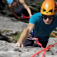 Un grimpeur concentré, casque orange, s'engage dans une voie d'escalade 64. Une corde rouge vif l'assure.