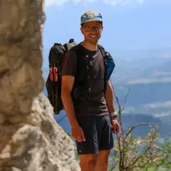 Jérémy Bigé, souriant, prêt pour l'aventure avec son sac à dos et ses bâtons de randonnée, dominant un paysage de montagnes.