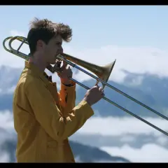 Victor Decamp Destivelle, un jeune homme aux cheveux ébouriffés, joue du trombone dans un paysage montagneux brumeux.