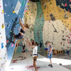 Une femme fait de l'escalade climb up sur un mur coloré, d'autres personnes observent ou se préparent.