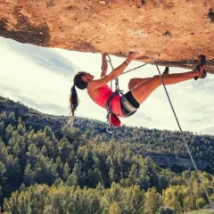 Une femme en pleine escalade extrême, suspendue sous un surplomb rocheux, avec une forêt verdoyante en arrière-plan.