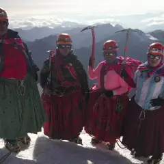 Quatre cholitas escaladoras, casques et piolets, posent fièrement sur un sommet enneigé, leurs jupes colorées flottant au vent.
