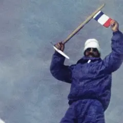 Les mains de Maurice Herzog brandissent un drapeau français au sommet d'une montagne enneigée, symbole d'une victoire alpine.