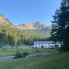 Vue panoramique de la **selva de Oza** avec une bâtisse blanche, des montagnes imposantes et une forêt dense sous un ciel bleu clair.