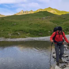 Un randonneur traverse un ruisseau en utilisant des bâtons de marche. Le soleil éclaire les montagnes verdoyantes en arrière-plan.