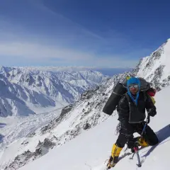 Daniele Nardi, alpiniste, en pleine ascension sur une pente enneigée, avec un sac à dos imposant et des crampons. Le paysage montagneux est spectaculaire.