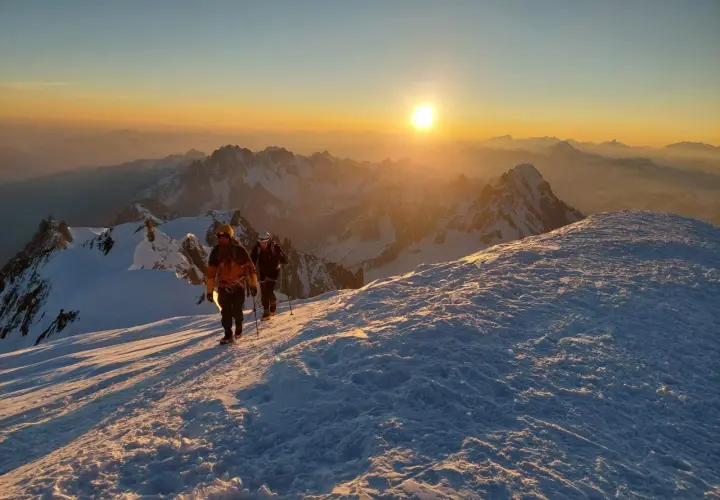 Deux alpinistes progressent sur un sentier enneigé au lever du soleil, dominant un paysage de montagnes spectaculaire. Le sommet montagneux est baigné d'une lumière dorée.