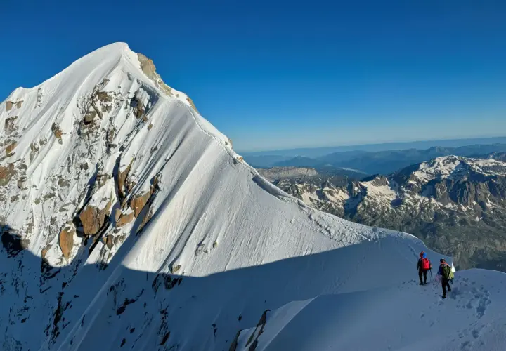 Deux alpinistes escaladent la crête enneigée de l'Aiguille Verte, sous un ciel bleu intense. Le soleil projette de longues ombres sur la montagne.