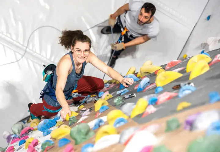 Une femme souriante en tenue d'escalade grimpe un mur coloré, assurée par un homme en arrière-plan.