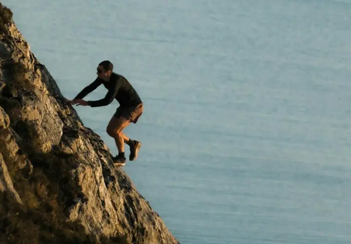 Kilian Jornet escalade une paroi rocheuse abrupte, dominant un plan d'eau calme, rappelant l'esprit d'aventure dans les Pyrénées.