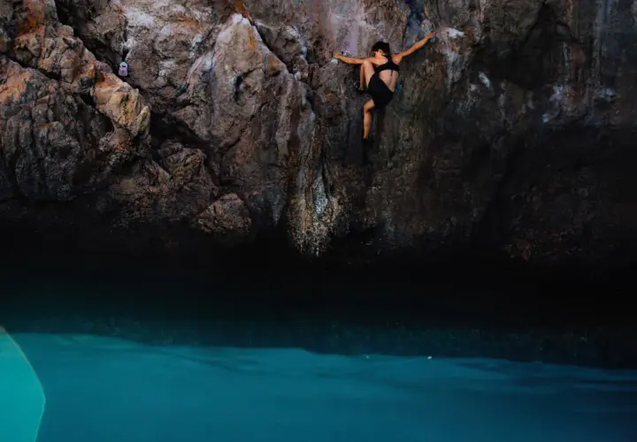 Une femme pratique l'escalade outdoor au-dessus d'une eau turquoise. Elle s'agrippe à la paroi rocheuse, concentrée sur sa prochaine prise.