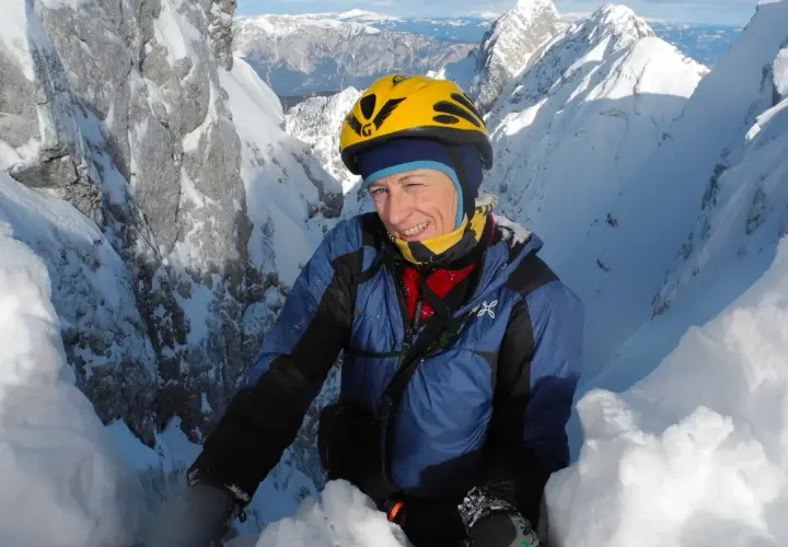 Une alpiniste souriante, Nives Meroi, en plein effort sur une crête enneigée, casque jaune, montagnes majestueuses en arrière-plan.