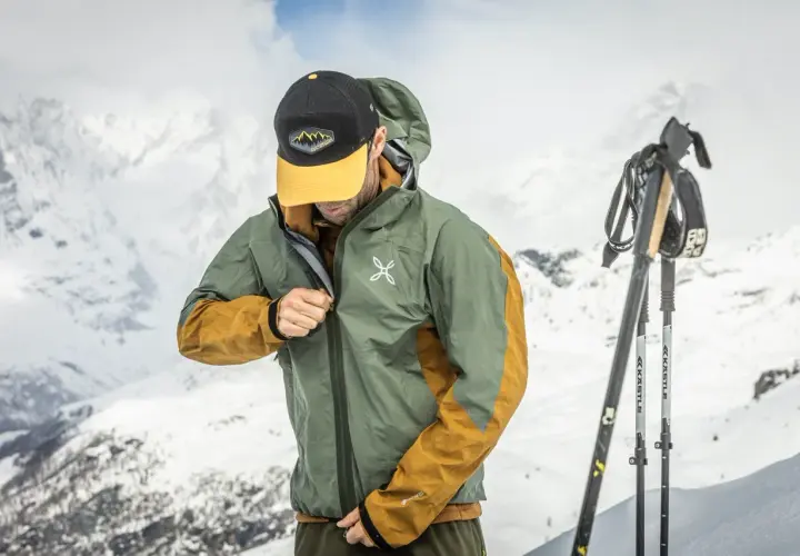 Un skieur ajuste sa veste technique, peut-être pour comparer le confort entre Primaloft vs Gore-Tex, avec des montagnes enneigées en arrière-plan.