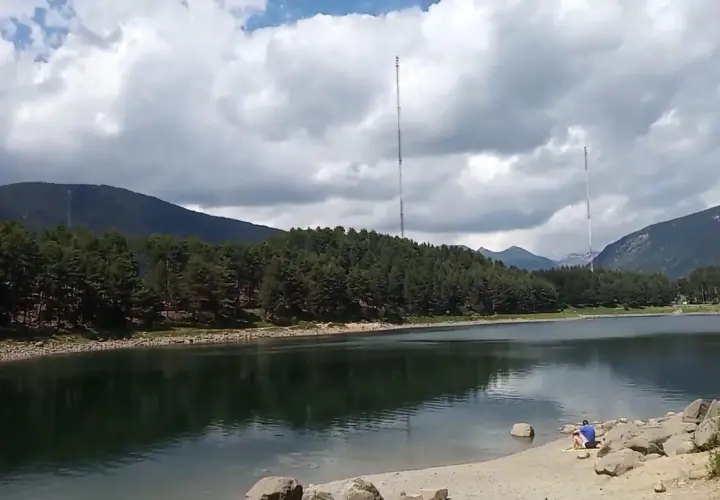 Vue paisible du lac d'Engolasters, entouré de forêts de pins et de montagnes. Deux tours de transmission se dressent à l'horizon.