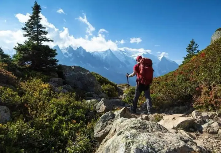Un randonneur solo, sac à dos rouge, bâtons de marche, admire les montagnes majestueuses sous un ciel bleu éclatant.