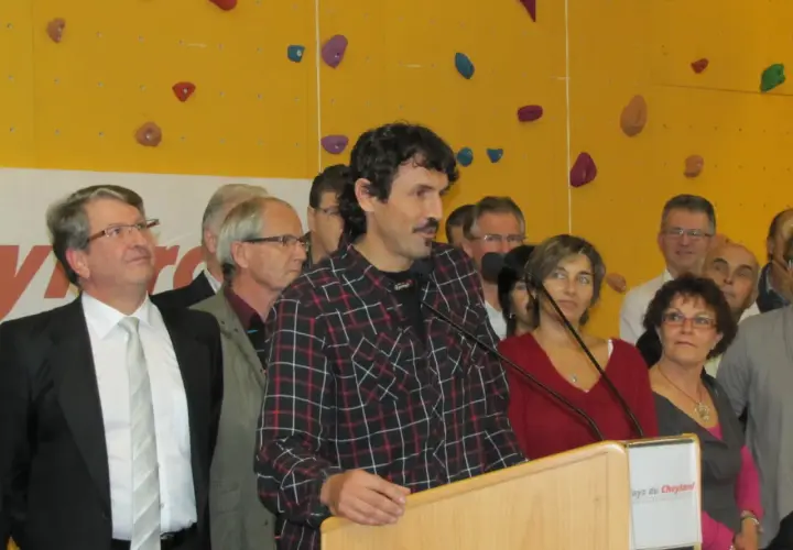 Antoine Cayrol, orateur charismatique, s'exprime devant un mur d'escalade coloré, entouré d'une foule attentive.