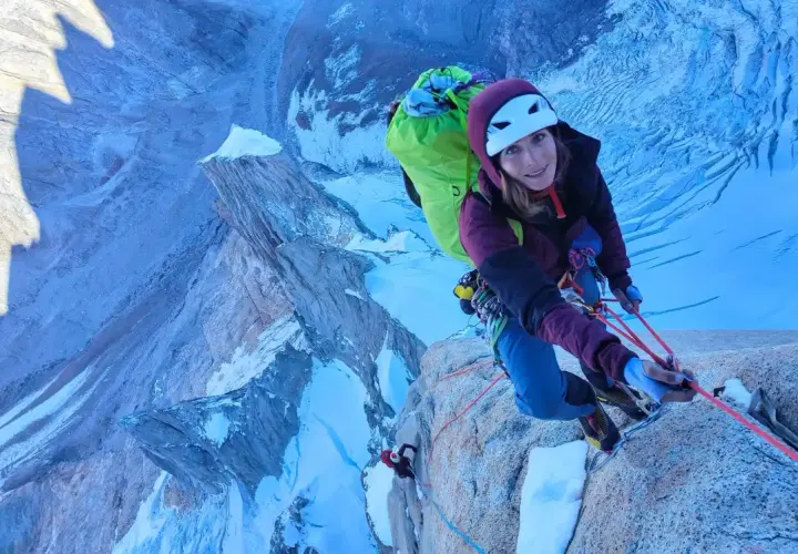 Lise Billon, casque blanc, grimpeuse souriante, est en pleine ascension sur une paroi rocheuse glacée, avec un sac à dos vert vif.