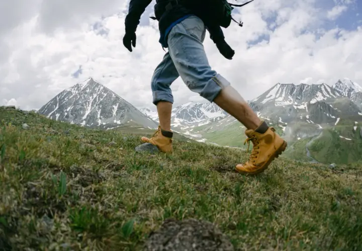 Un randonneur en bottes jaunes gravit une pente herbeuse, les montagnes enneigées en toile de fond. Une belle rando montagne.