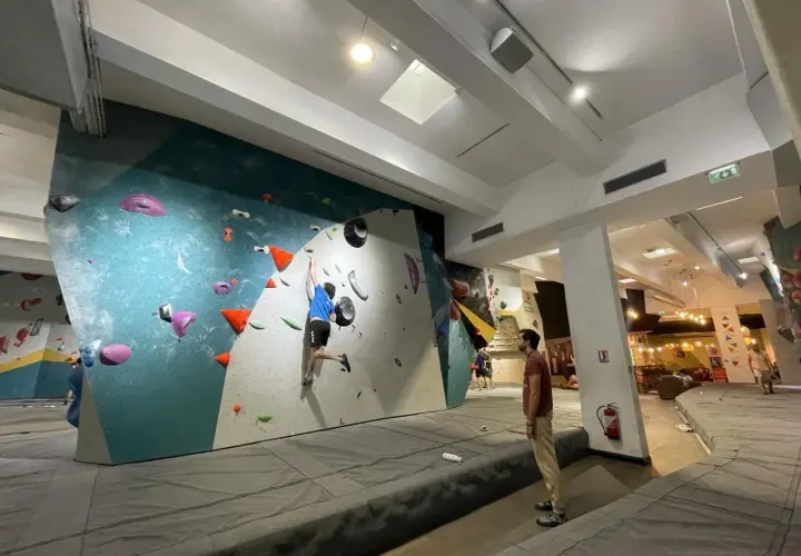 Un jeune homme en bleu pratique l'escalade sur un mur coloré, un autre spectateur l'observe. L'ambiance est propice à l'escalade nation.