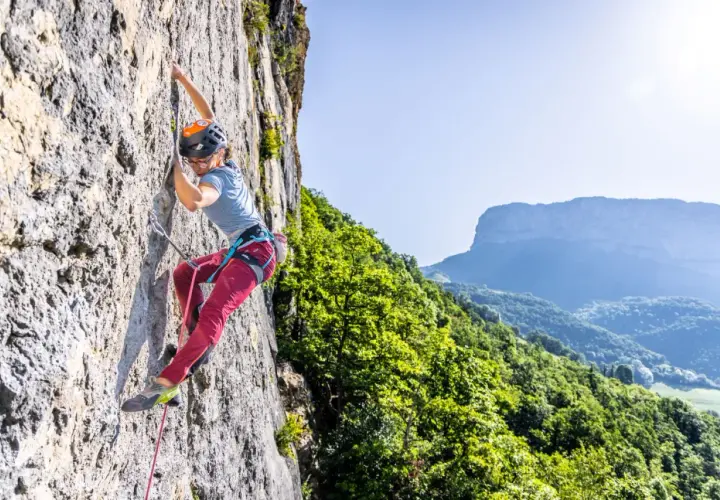 Une grimpeuse évolue sur une paroi rocheuse, profitant d'une journée ensoleillée dans ce parc d'escalade naturel. Le paysage montagneux verdoyant offre un panorama spectaculaire.