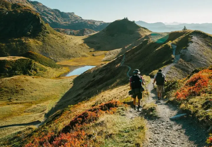 Deux randonneurs avancent sur un sentier de montagne, profitant des paysages spectaculaires. Les couleurs d'automne ajoutent une touche vibrante à la scène, tandis que les fiz s'élèvent au loin.