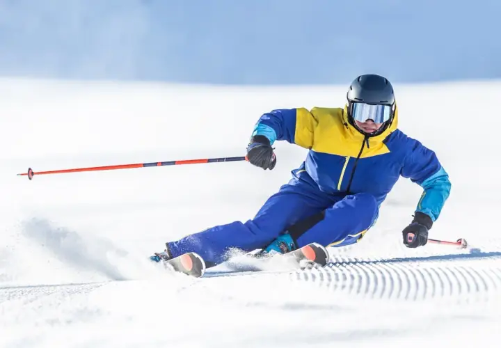Un skieur en combinaison jaune et bleue vire sur la neige, ses skis paraboliques taillant la piste.