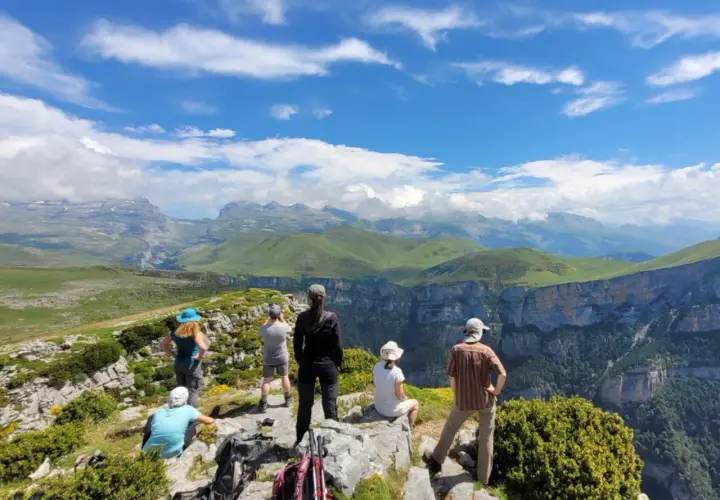 Randonneurs contemplant le paysage spectaculaire du **parque nacional de Ordesa y Monte Perdido**, avec ses falaises abruptes et ses montagnes verdoyantes sous un ciel bleu.