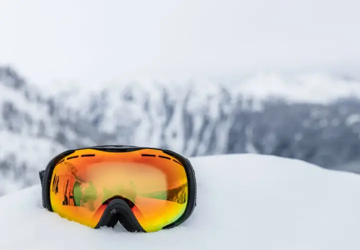 Masque de ski orange posé dans la neige, reflétant des skieurs. Indispensable pour savoir quel masque de ski choisir.