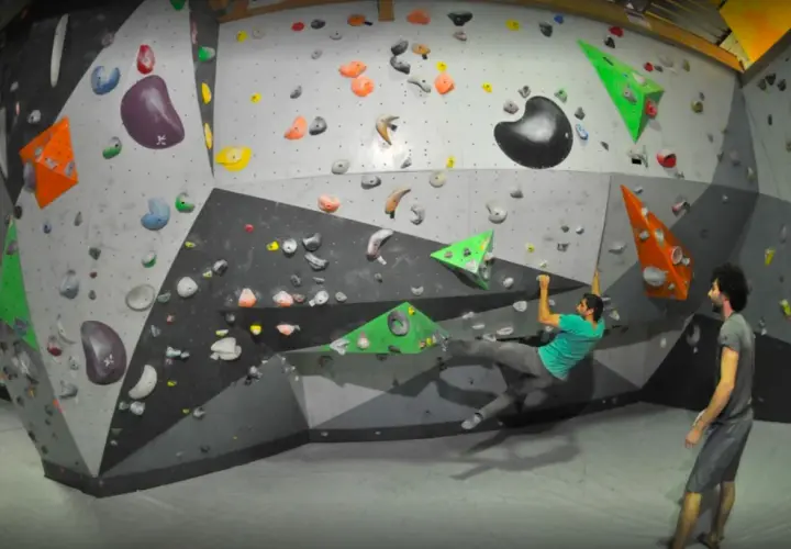 Un homme en t-shirt vert fait de l'escalade pertuis sur un mur coloré, un autre homme le regarde.