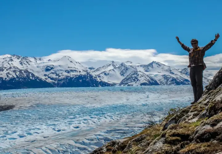 Un homme bras levés devant un glacier immense et des montagnes enneigées de la Patagonie.