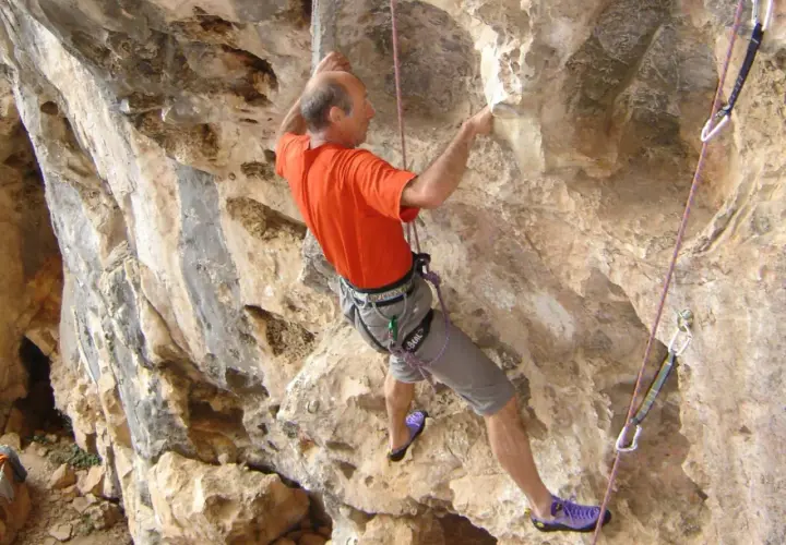 Un homme en t-shirt orange et short gris fait de l'escalade sur une paroi rocheuse. Il est assuré par une corde et semble atteindre un point élevé, un vrai **grimp azur**.