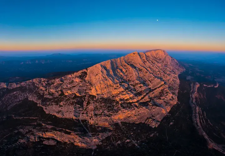 Le soleil couchant illumine la montagne Sainte-Victoire, offrant un spectacle grandiose pour les amateurs d'escalade. La lune veille sur ce paysage rocheux.